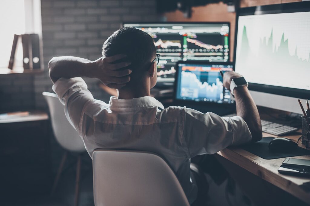 become a trader - Rear view of young man in casual wear holding hand on the back of the head and working while sitting at the desk in creative office